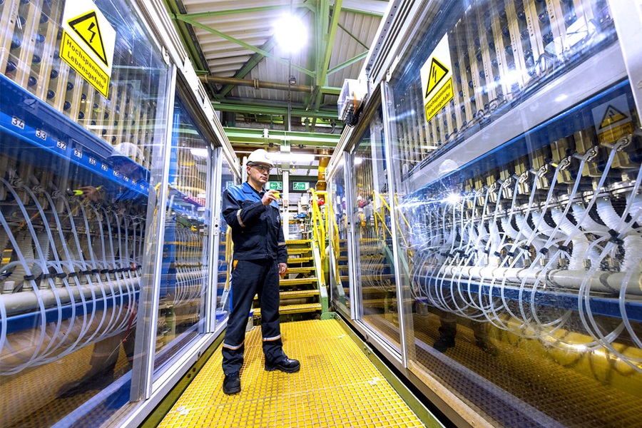 An employee wearing working clothes is inspecting a room full of glass cupboards which contain lots of tubes and cables.