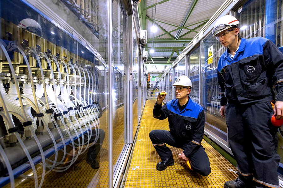 Two employees wearing protective glasses and working clothes are inside the room of a power station and inspecting tubes and cables protected by glass doors.