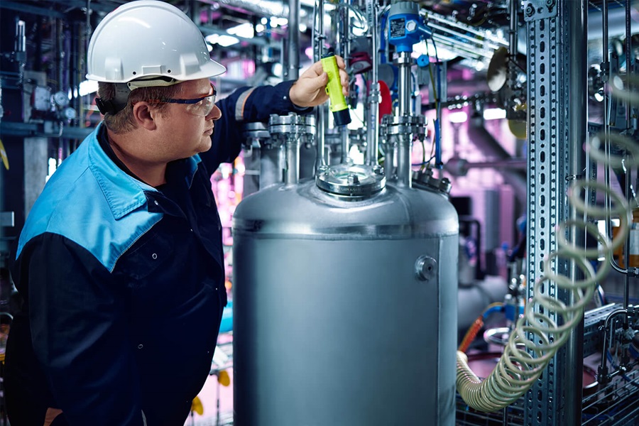An employee in a factory is wearing protective glasses, a helmet and working clothes and is holding a yellow flashlight in order to inspect a machine