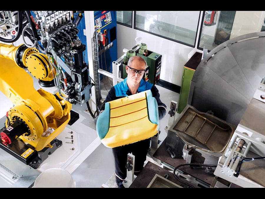 Factory employee wearing protective glasses and working clothes is standing next to a machine and inspecting a part of a vehicle seat.