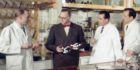 Hermann Schnell holds an object in his hand in a laboratory and talks to three men in lab coats.
