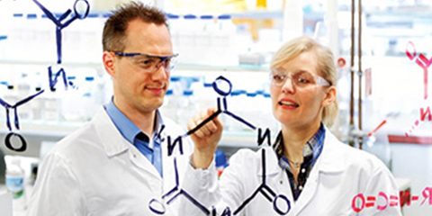 Two lab technicians write chemical formulas on a blackboard.