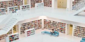 Multilevel library with many books, white floors and shelves, seating areas, and a staircase.