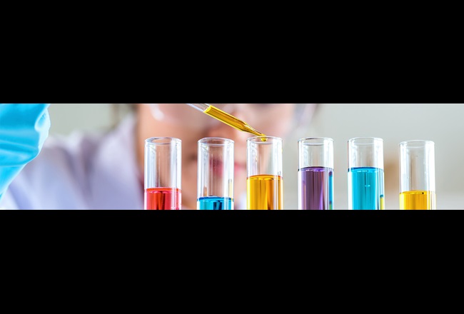 A woman is filling a liquid into a test tube using a pipette. Several test tubes with colorful liquids are in front of her.